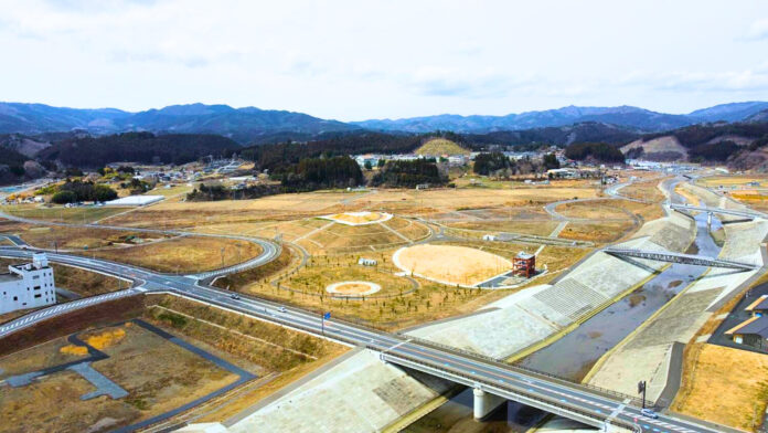 広い平地に円形の区画がいくつも描かれ、右側には直線的な川とその両側に高い堤防が見え、奥には山並みと小さな集落が広がっている復興中の町並みの昼間の風景を高台から定点カメラで撮影した写真