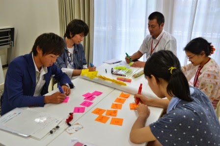 5人の参加者がカラフルな付箋にペンで意見を書いているグループワークの様子の写真