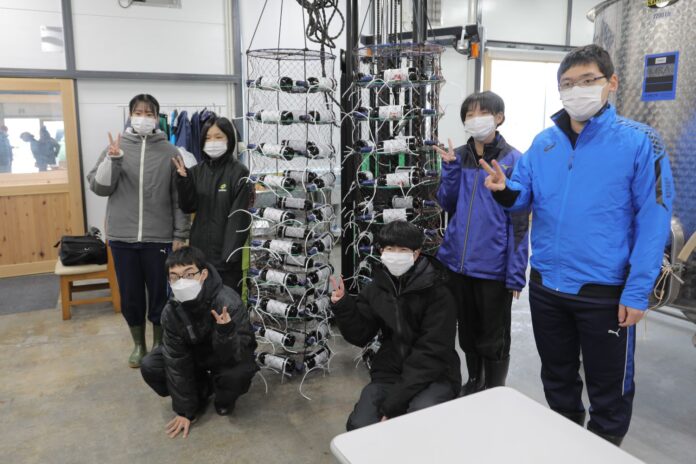 6人の高校生たちが施設内で天井から吊り下げられたたくさんのワインボトルが入った海中熟成用のかごの前でピースサインをしている写真