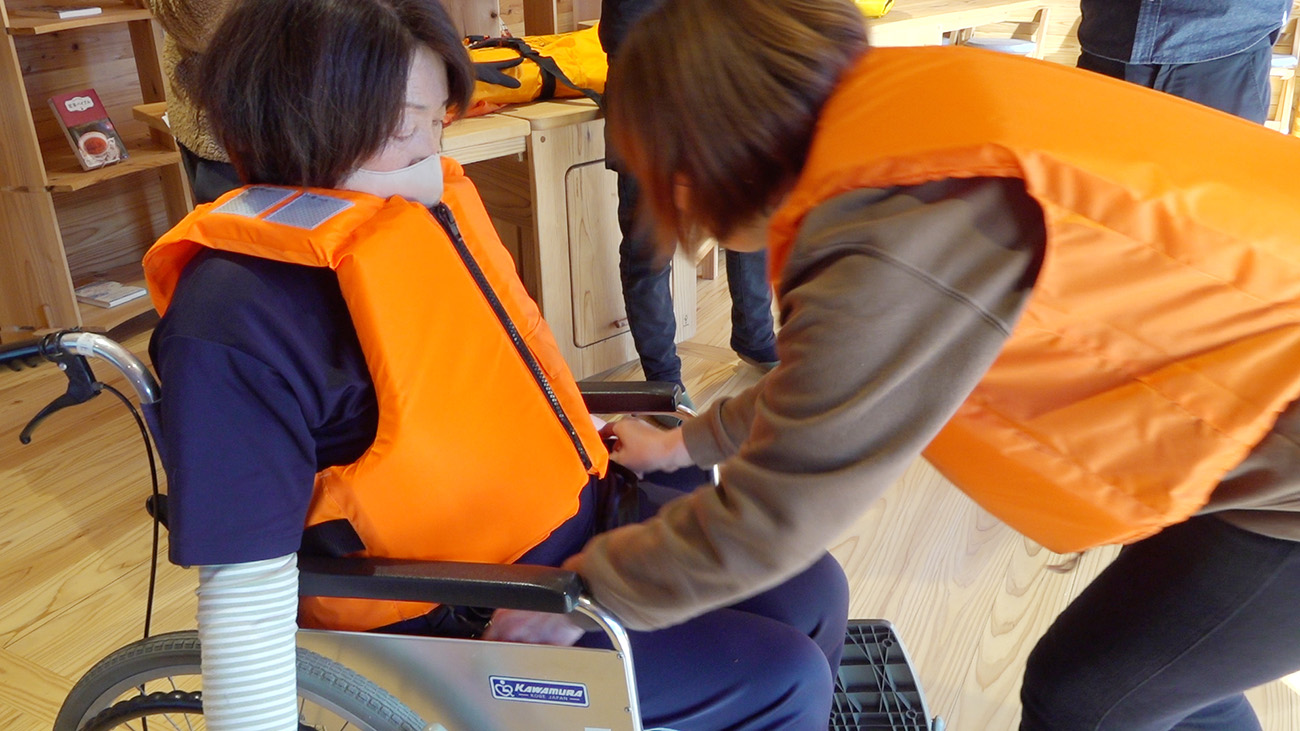木の床と壁に囲まれた室内で、車椅子に座った女性がオレンジ色のライフジャケットを着ており、もう一人がそれを調整している様子の写真