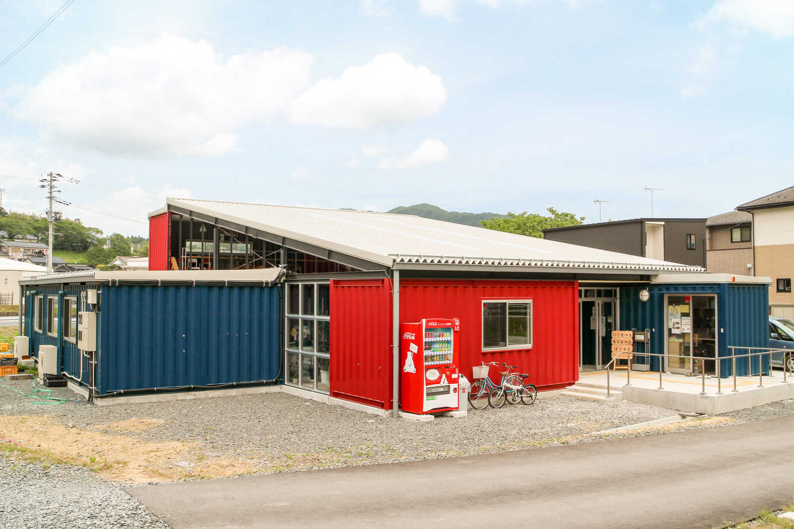 赤と青のコンテナでできた平屋の建物で、壁沿いに自動販売機と自転車が置かれている、晴谷驛（はればれー）の外観写真