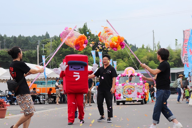 中央に平仮名で「う」と書かれた赤いポストの形の着ぐるみキャラクターと黒い服を着た男性が、紙吹雪が舞う中で行進している様子の写真