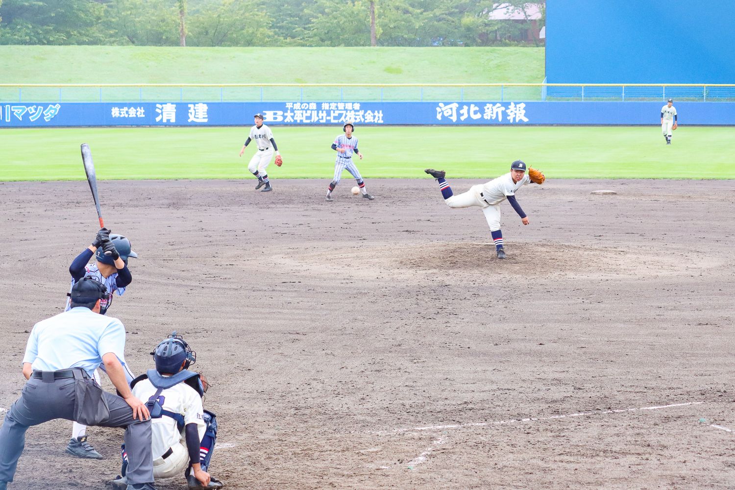 平成の森・しおかぜ球場で、ピッチャーがボールを投げ、バッターがスイングしようとしている瞬間を捉えた、高校野球の県大会の試合中の一場面の写真