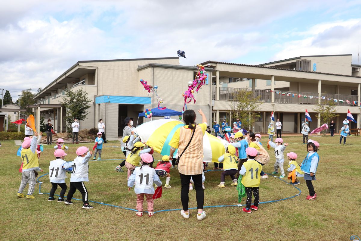芝生の広場で子どもたちがパラバルーンを広げ、周囲の園児や先生たちも掛け声に合わせて一緒にダンスを披露している様子の写真