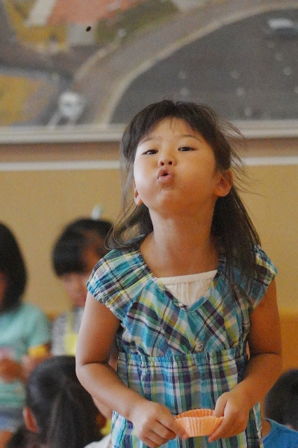 チェック柄の服を着た女の子が一生懸命に口をとがらせてスイカの種を飛ばしている様子の写真