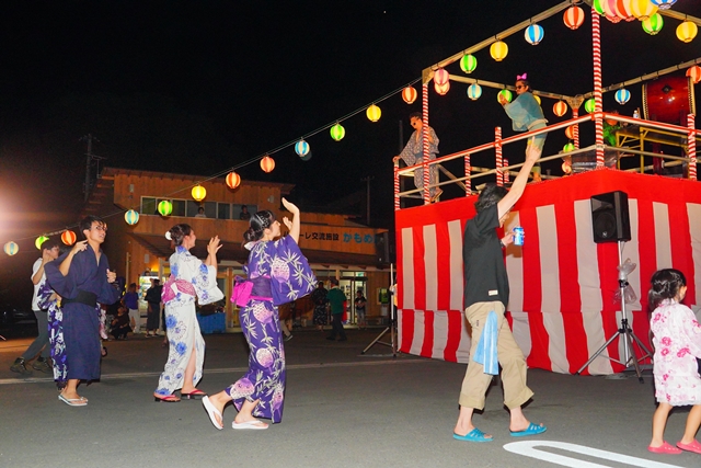 提灯が飾られたやぐらの前で、浴衣を着た人々が音楽に合わせて盆踊りをしている夜の祭りの様子の写真