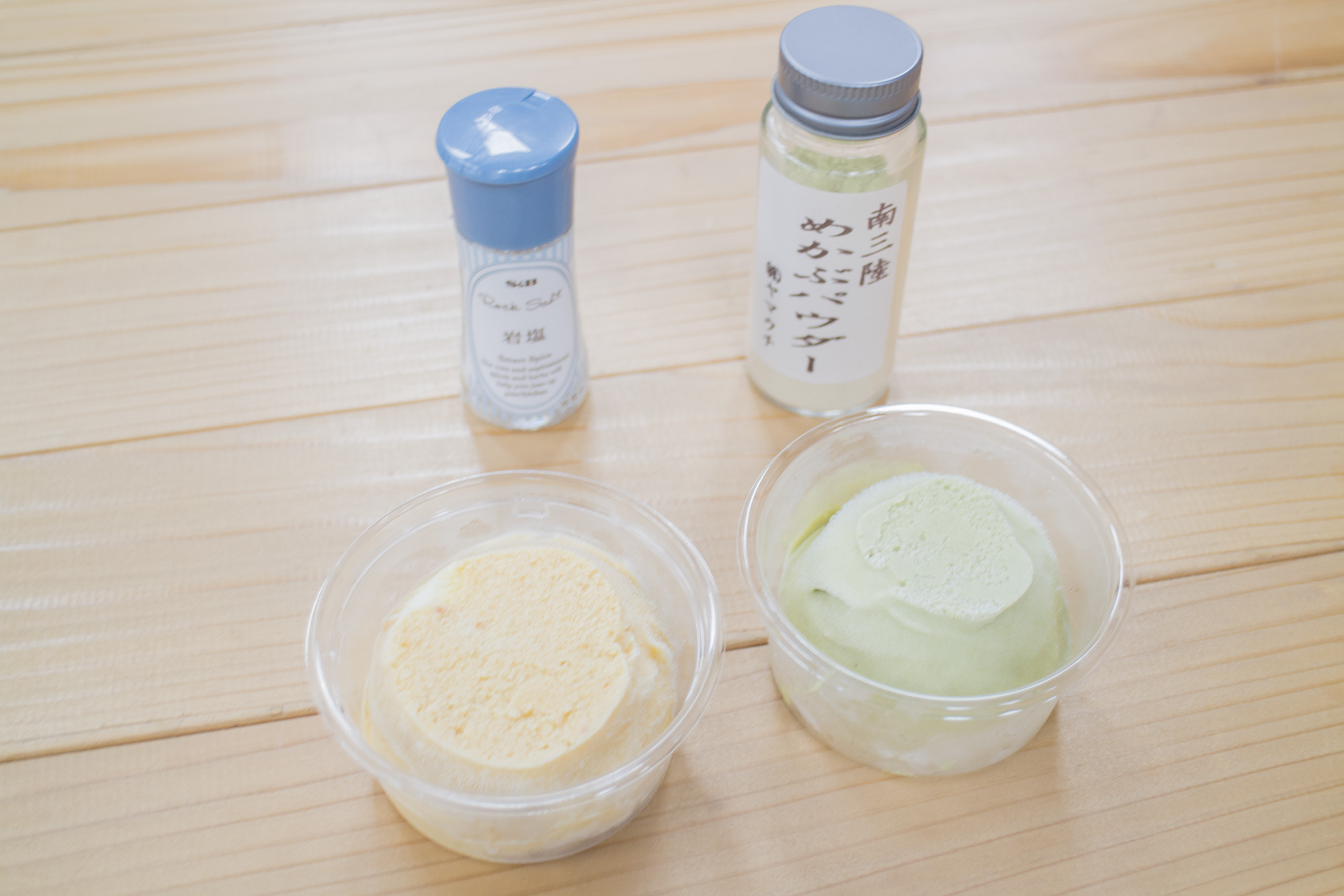 木のテーブルの上に、透明のカップに入った、白と薄い緑の2種類のご当地アイスと、岩塩、「南三陸めかぶパウダー」の瓶が並べて置かれている写真