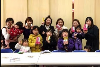 後列に大人の女性が7人前列に子どもが5人並び室内のテーブルの前で飲み物の缶やパッケージを手に持ちカメラに向かって明るい笑顔をしている様子を写した写真