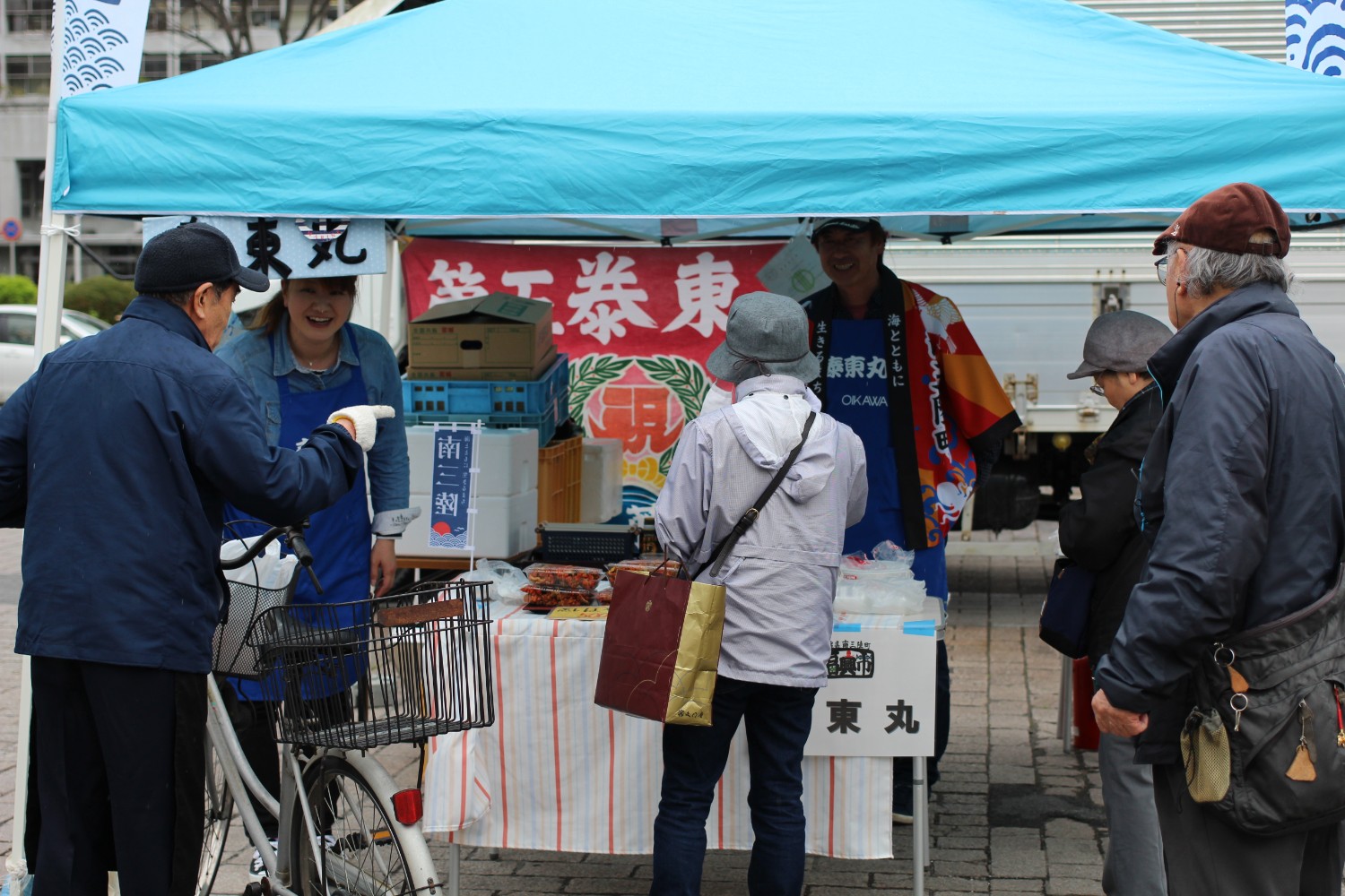 青いテントの下のブースの前に買い物客が集まり、法被やエプロンを着たスタッフたちが笑顔で対応している写真