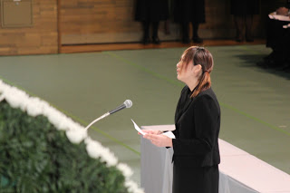 遺族代表の佐藤綾美さんがマイクの前に立ち、祭壇に向かって慰霊の言葉を述べている場面を写した写真
