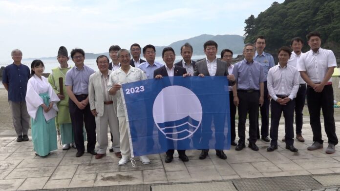 後方には山や海が広がる穏やかな風景の中で海辺の遊歩道のような場所で、神主や巫女を含む約20人の中心のスーツ姿の男性たちが「2023」と書かれた青い旗を持ち、集合写真を撮っている写真