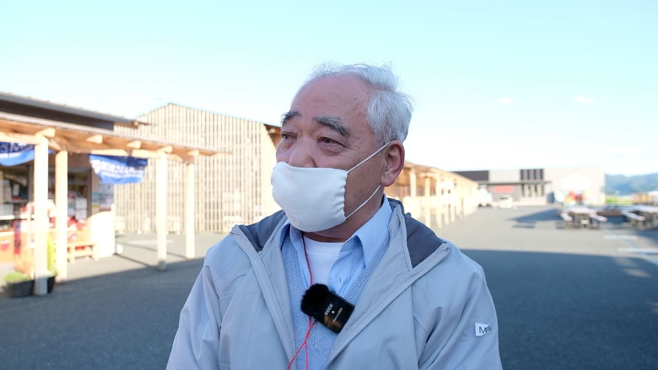 マスクをつけた伊東正和さんが屋外の商業施設前でインタビューを受けており、静かな雰囲気の中で復興市の背景について語っているインタビューの写真