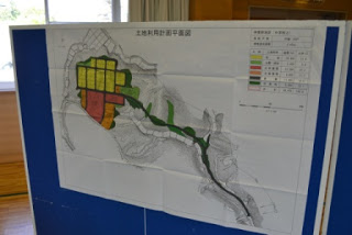 会議会場の一角に白地の地図に赤や緑などで色分けされた中学校上団地の土地利用計画図が掲示されている様子の写真