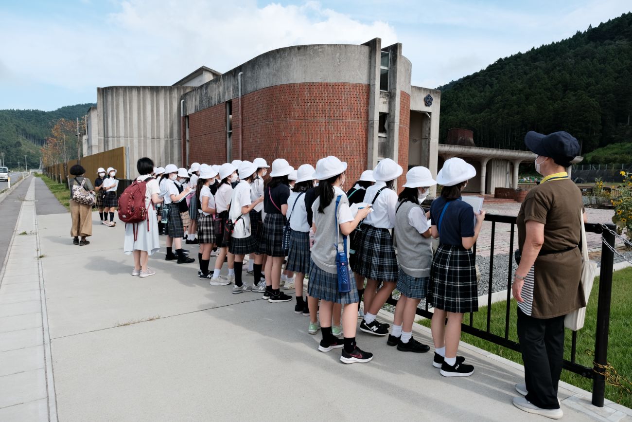 白い帽子と制服姿の女子学生たちが、震災遺構と思われる建物を見学しながらメモを取っている様子の写真
