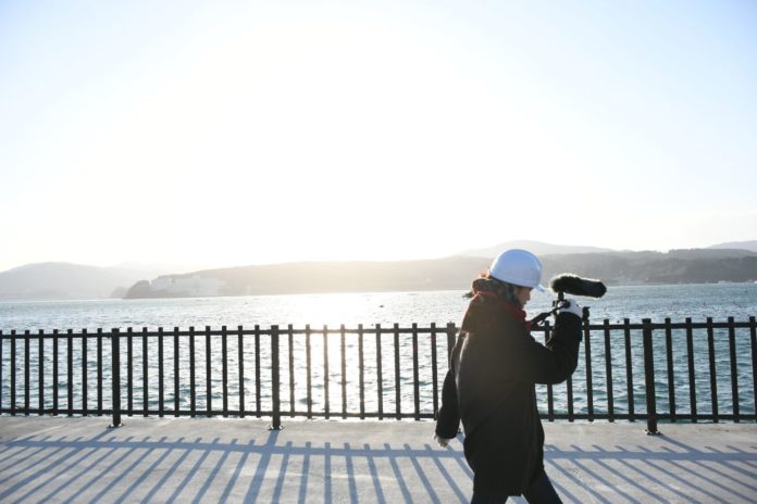 白いヘルメットをかぶった女性がマイク付き録音機材を持ち、太陽に照らされる海沿いの舗装された道を歩いている写真