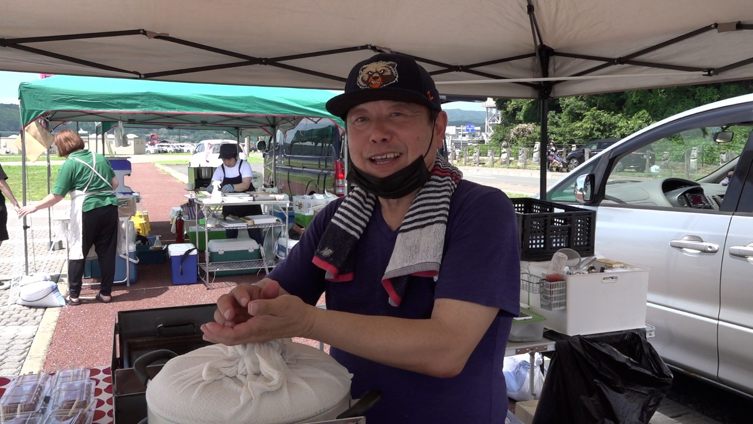後ろには調理中の女性たちと車が並ぶ野外屋台テントの下で、笑顔の男性が肩にはタオルをかけ両手を合わせて接客の準備をしている写真