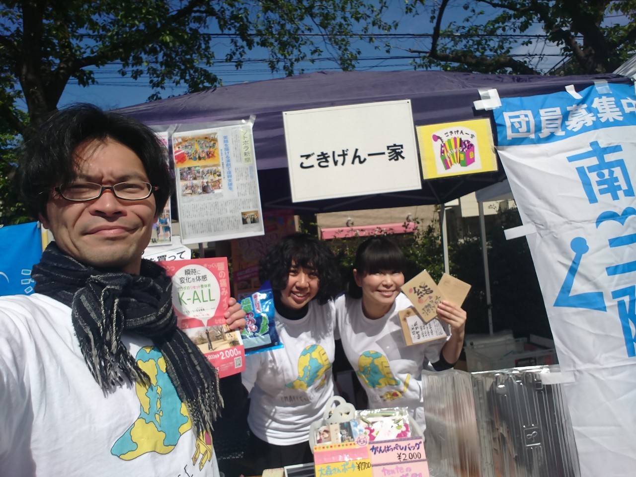 「ごきげん一家」と書かれた屋外の出店ブースの前で、お揃いの白いTシャツを着た3人の男女が手に商品を持ち笑顔で写っている写真