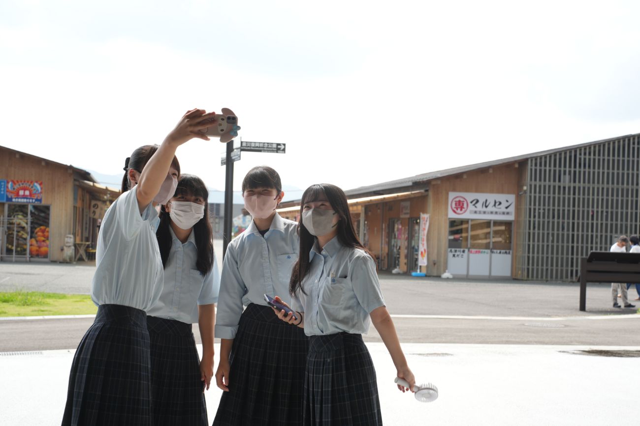 制服を着た高校生4人が屋外でスマートフォンを使って自撮りをしている様子の写真