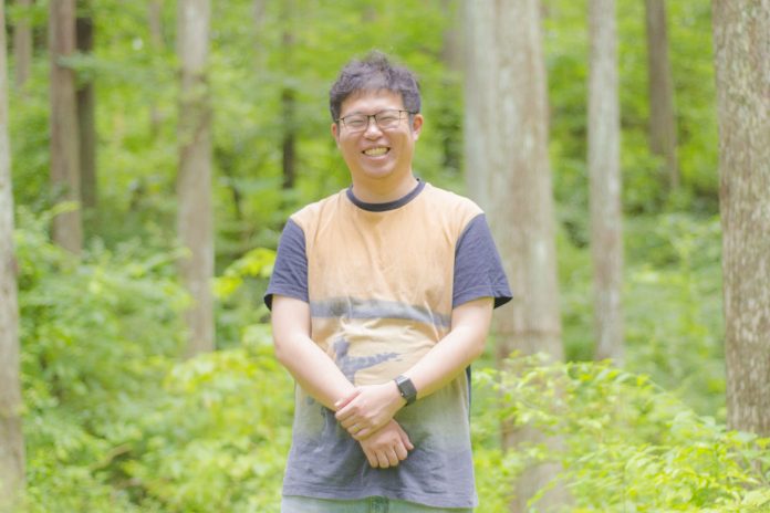 青々とした木々が生えている森の中で、両手を体の前で組み笑顔で立っている佐藤太一さんの写真