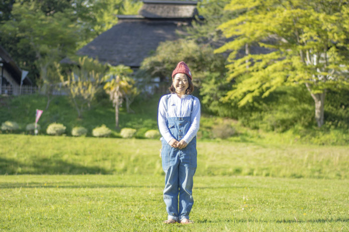 背景に茅葺き屋根の日本家屋と新緑が広がる草原の中で、赤いニット帽とデニムのオーバーオールを着た中島綾子さんが正面を向いて笑顔で立っている写真