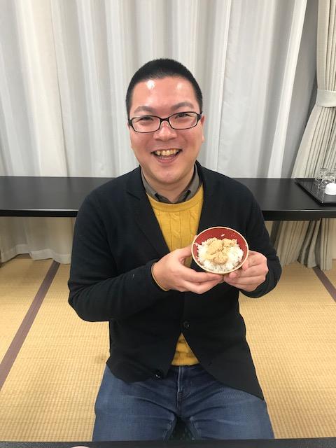 畳の部屋で笑顔を見せながら、白ご飯の上に鱈のタラコ煮がのった器を両手で持っている佐藤さんの写真