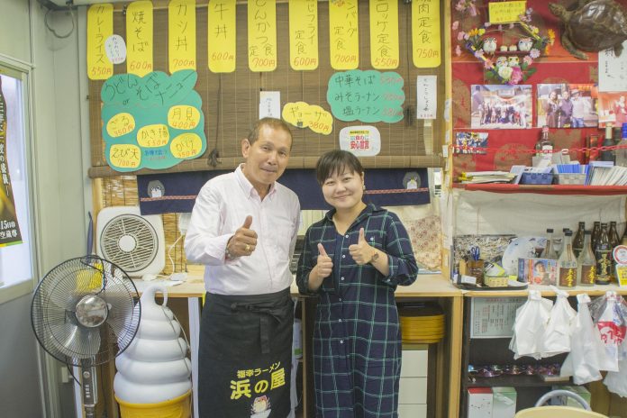 黄色い紙に書かれたメニュー札が並んだ食堂のカウンター前で、白いシャツに黒いエプロン姿の高橋さんと、女性リポーターが、笑顔で親指を立てたポーズて並んでいる写真