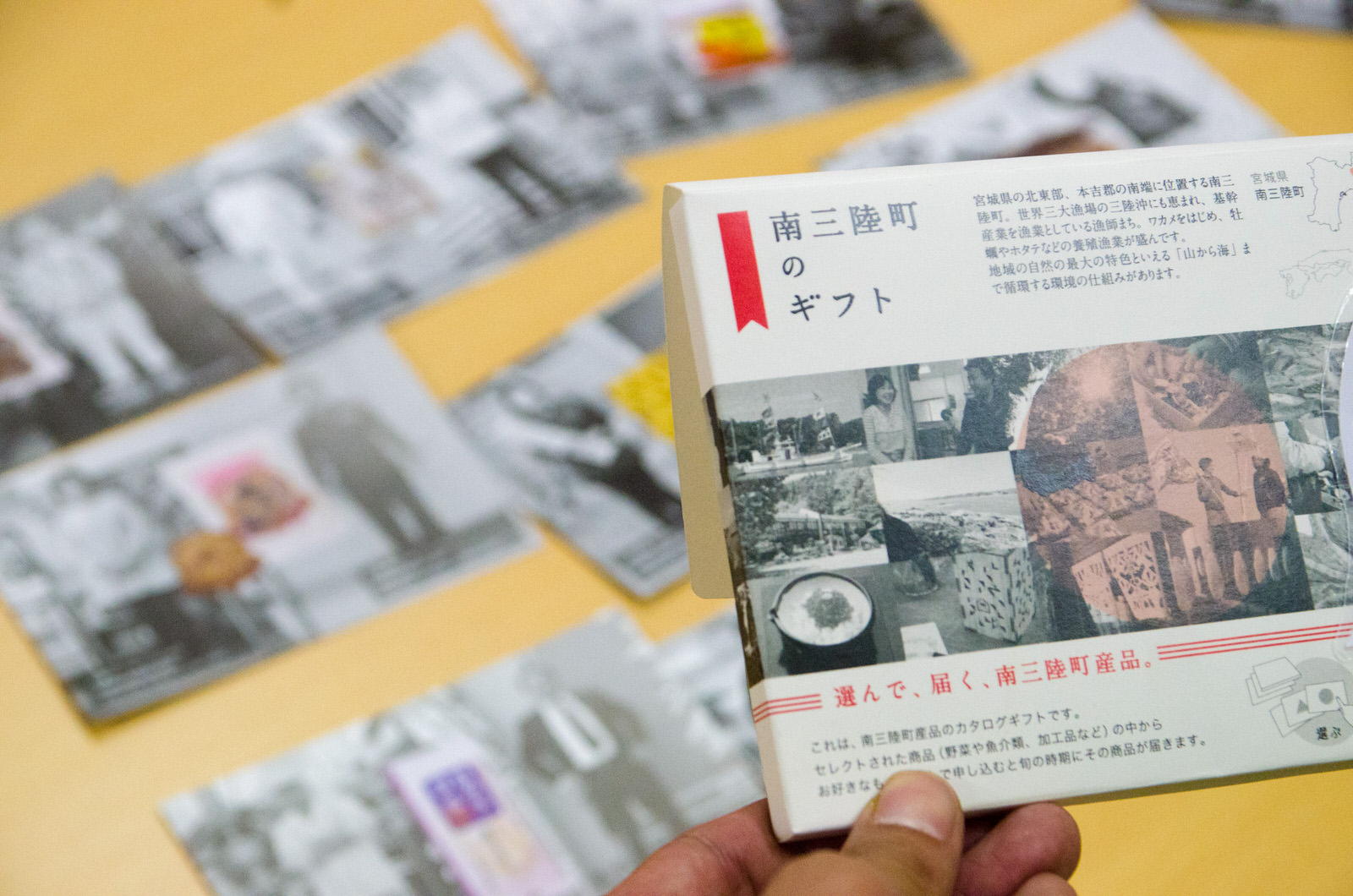 背景に黄色い机の上に置かれた沢山並べられたカードが、「南三陸町のギフト」と書かれたカタログギフトを持っている手越しに写っている写真