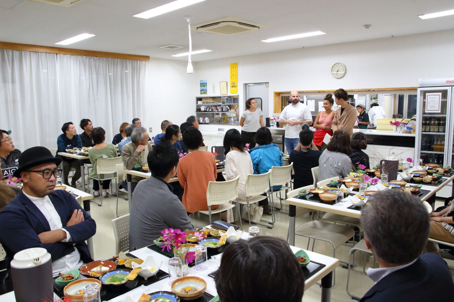 開発された料理が盛り付けられたお盆が置かれたテーブルの席に参加者が座り、3人のシェフと企画者の岩田さんの話を聞いている写真