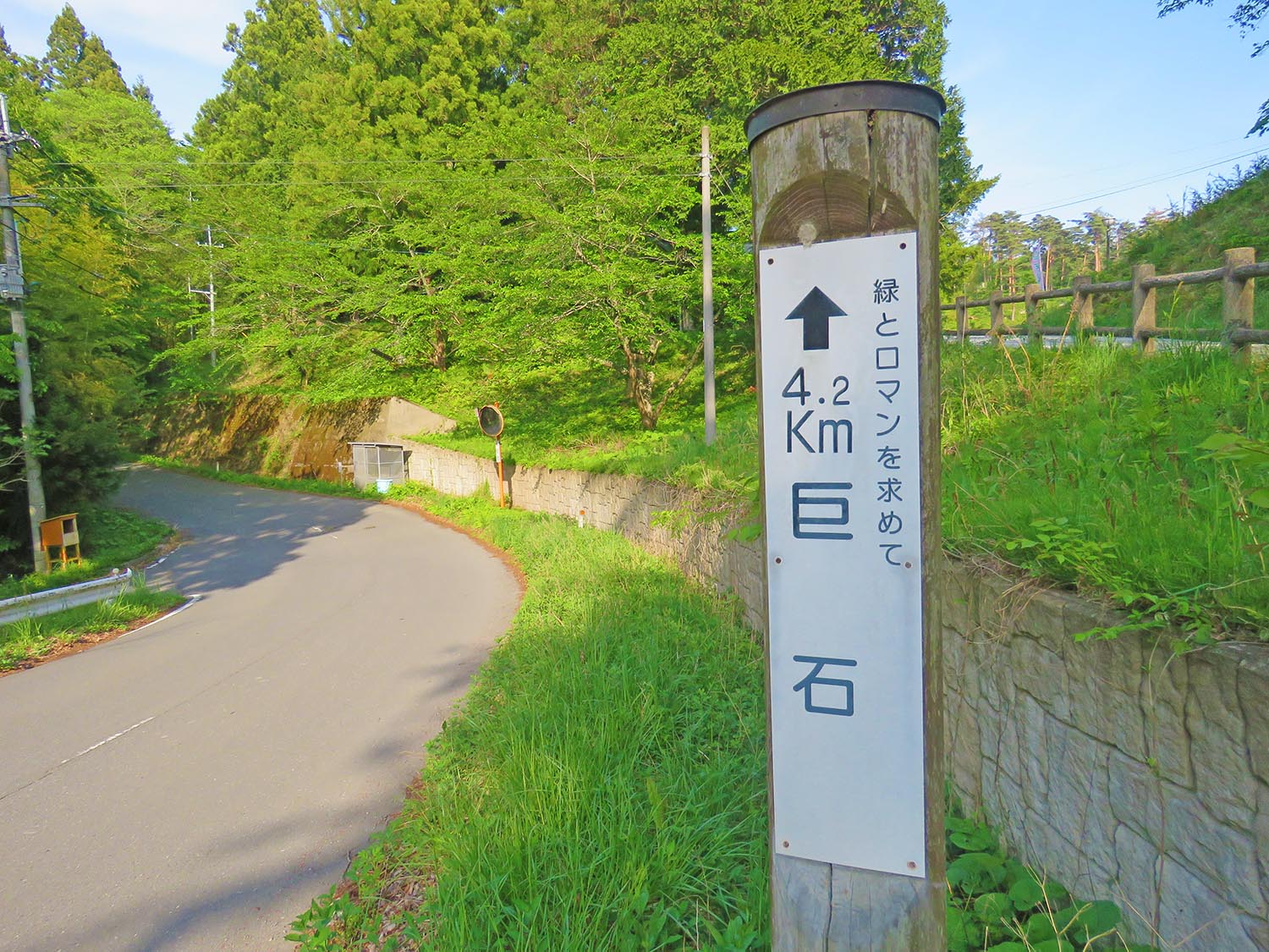 左側に向かってカーブする舗装された木々に囲まれた道の横に「緑とロマンを求めて 4.2キロメートル 巨石」と書かれた木製の柱が立っている写真