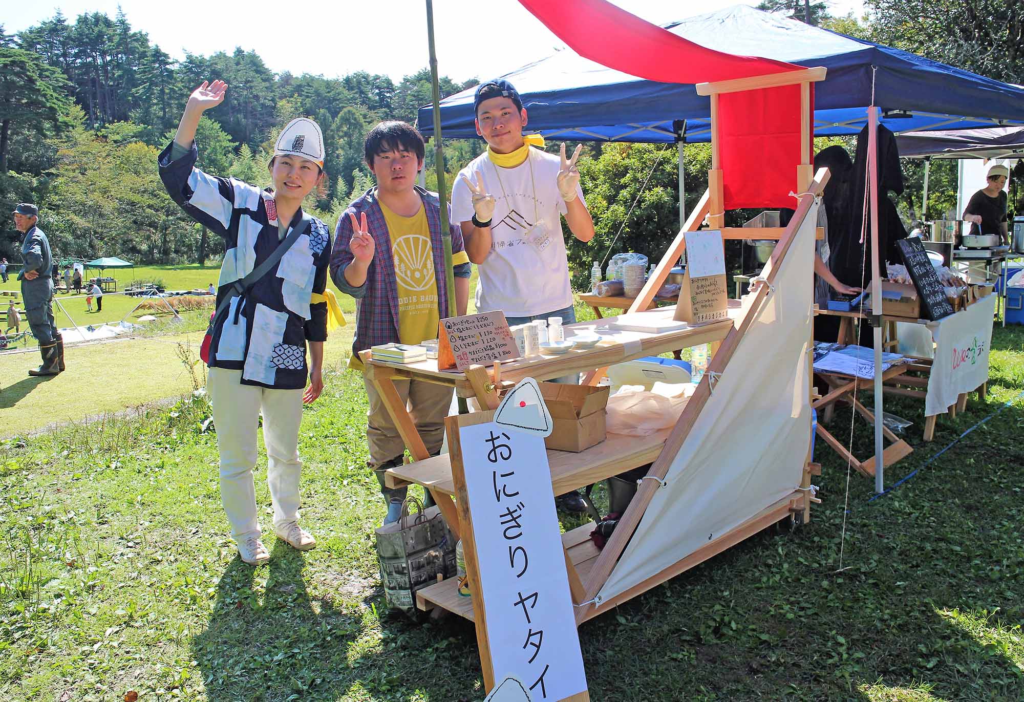 おにぎりヤタイと書かれた白い看板がある木製の屋台の前で、青空の下、3人のスタッフが笑顔でピースサインをしている、屋外イベントの様子の写真