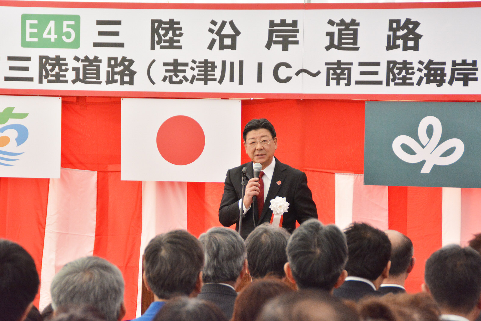 「E45 三陸沿岸道路」と書かれた横断幕と日の丸などの旗を背にマイクを持った佐藤町長が集まった人々の前で話をしている写真