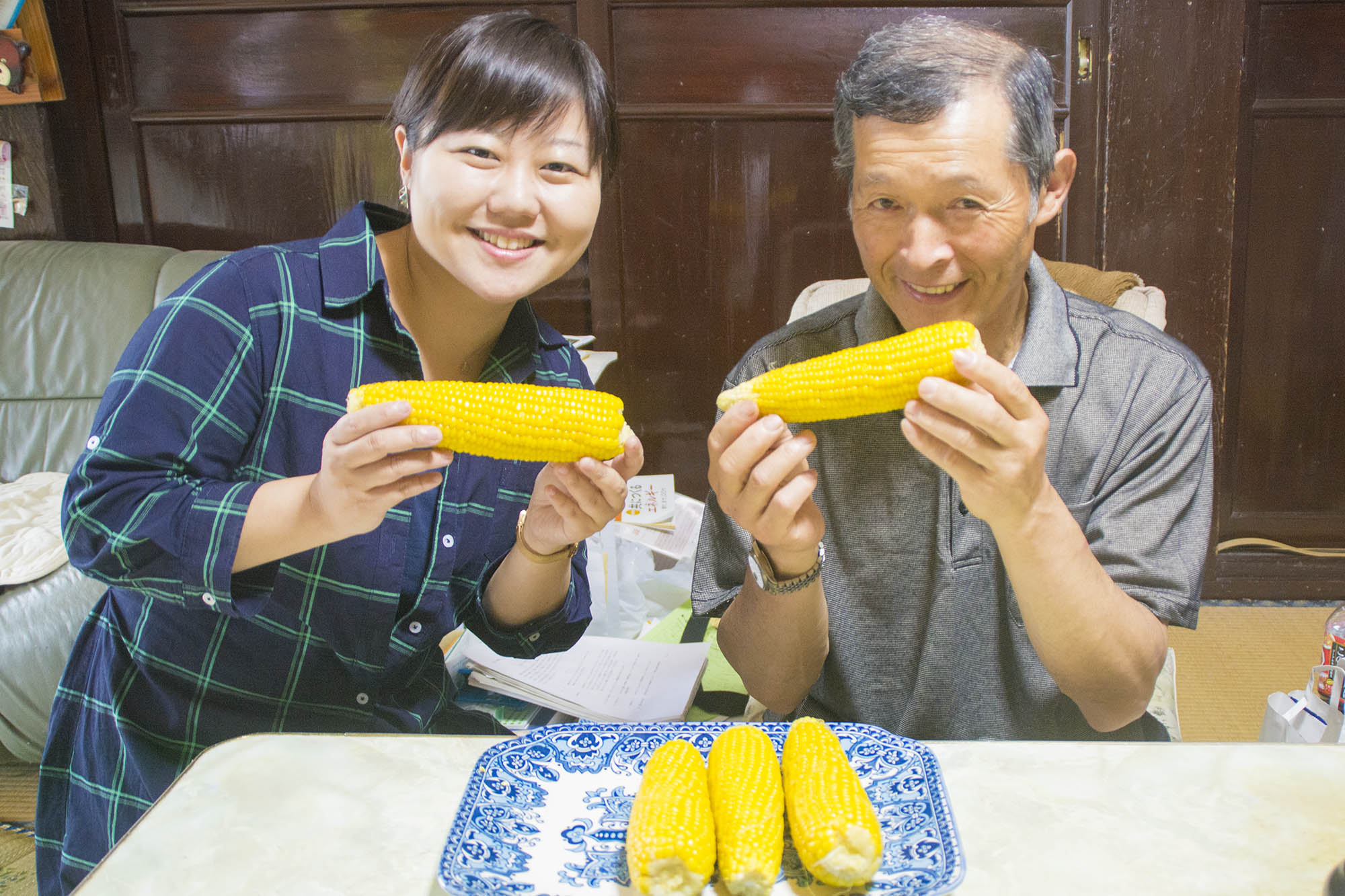 茹でトウモロコシが3本乗った皿がおかれたテーブルの後ろに座ったリポーターの女性と阿部さんが、それぞれ茹でたトウモロコシを手に笑顔でカメラを見ている写真