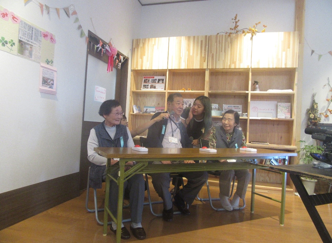 テーブルに座る高齢の女性二人と男性一人に向かって、女性スタッフが笑顔で説明しながら進行しているクイズの一場面の写真