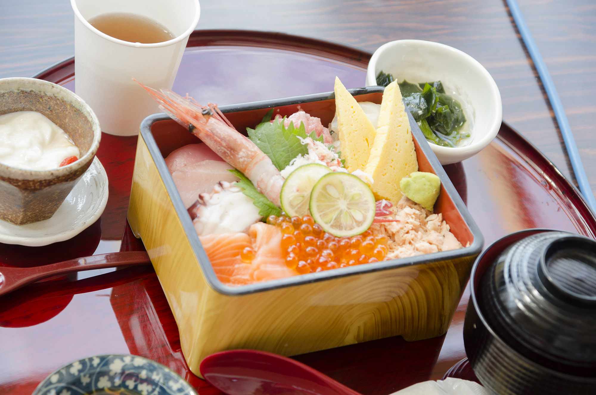 漆塗りのおぼんの上に、エビやイクラ、刺身、卵焼きなどの海鮮が盛られた色鮮やかな海鮮丼と味噌汁、小鉢などが並んでいる写真