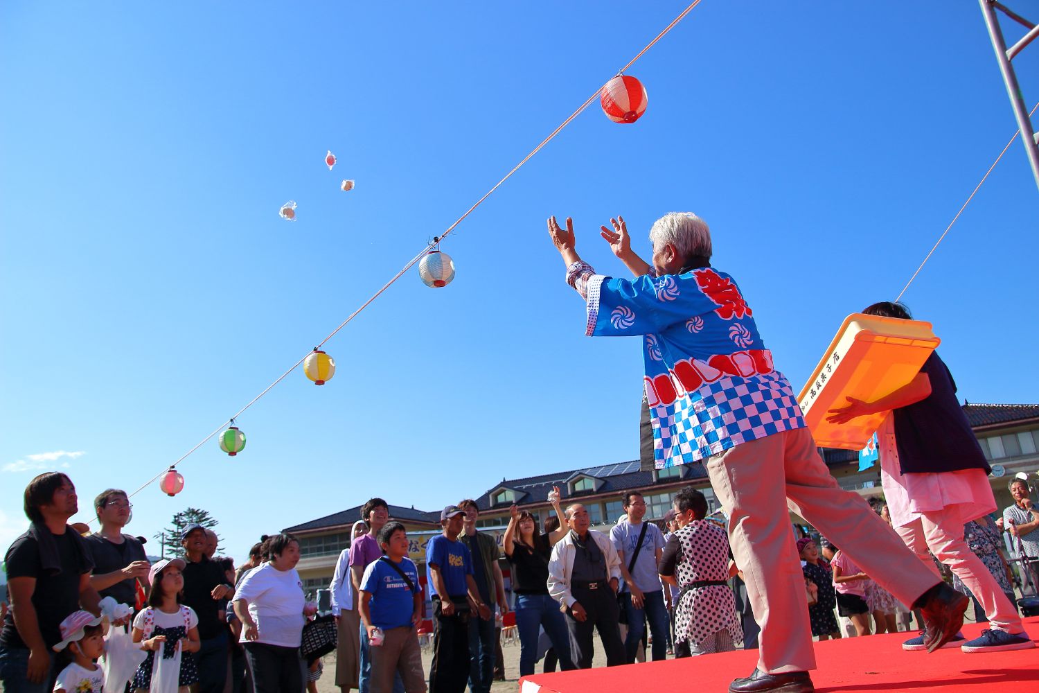 舞台の上で法被を着た白髪の人物が、空に向かって餅の入った袋を投げ、目の前にはそれを受け取ろうとする多くの観客が立っている写真