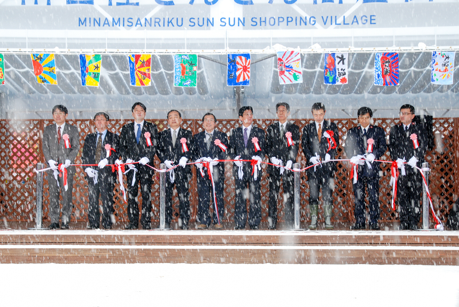 雪が降る中、手書きの大漁旗がたくさん飾られた壇上に上がったスーツ姿の男性たちがテープカットを行っている様子の写真