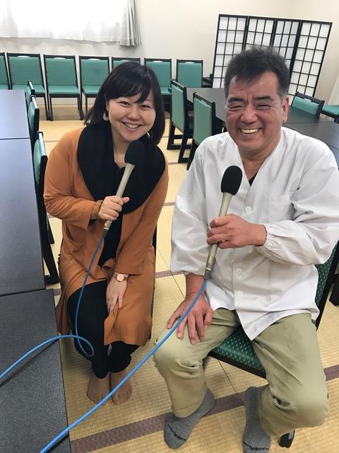 テーブルと椅子が並んだ畳敷きの室内で、マイクを持って笑顔で椅子に座っている女性リポーターと高橋さんの写真