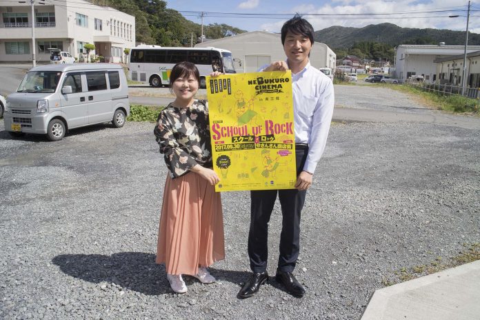 屋外の砂利の駐車場で、白いシャツを着た阿部将己さんと女性リポーターが、「SCHOOL OF ROCK」と書かれた黄色いポスターを一緒に持って立っている写真