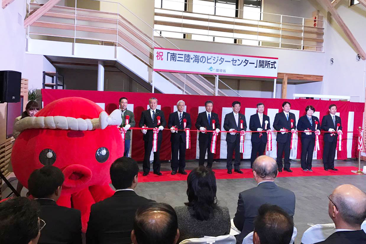「祝「南三陸・海のビジターセンター」開所式」と書かれた横断幕や紅白幕が貼られ、赤いオクトパス君の着ぐるみがいる会場で、レッドカーペットの上に横一列に並んだスーツ姿の関係者たちが、赤いリボンを持ってテープカットをしている様子の写真