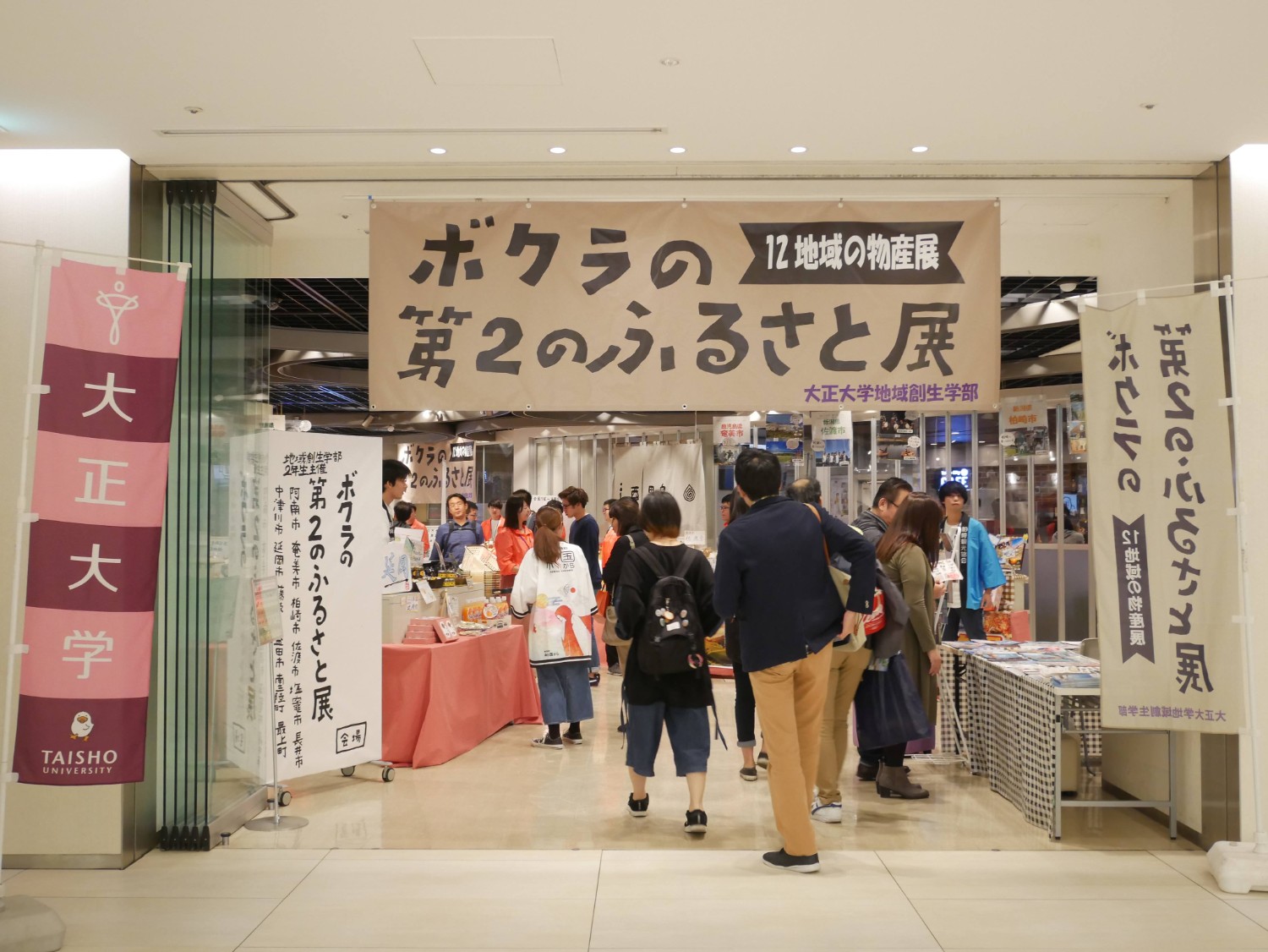 「ボクらの第2のふるさと展」と題された12地域の物産展が施設内で開催され、多くの人が展示品を見たり話をしたりしている様子を写した写真