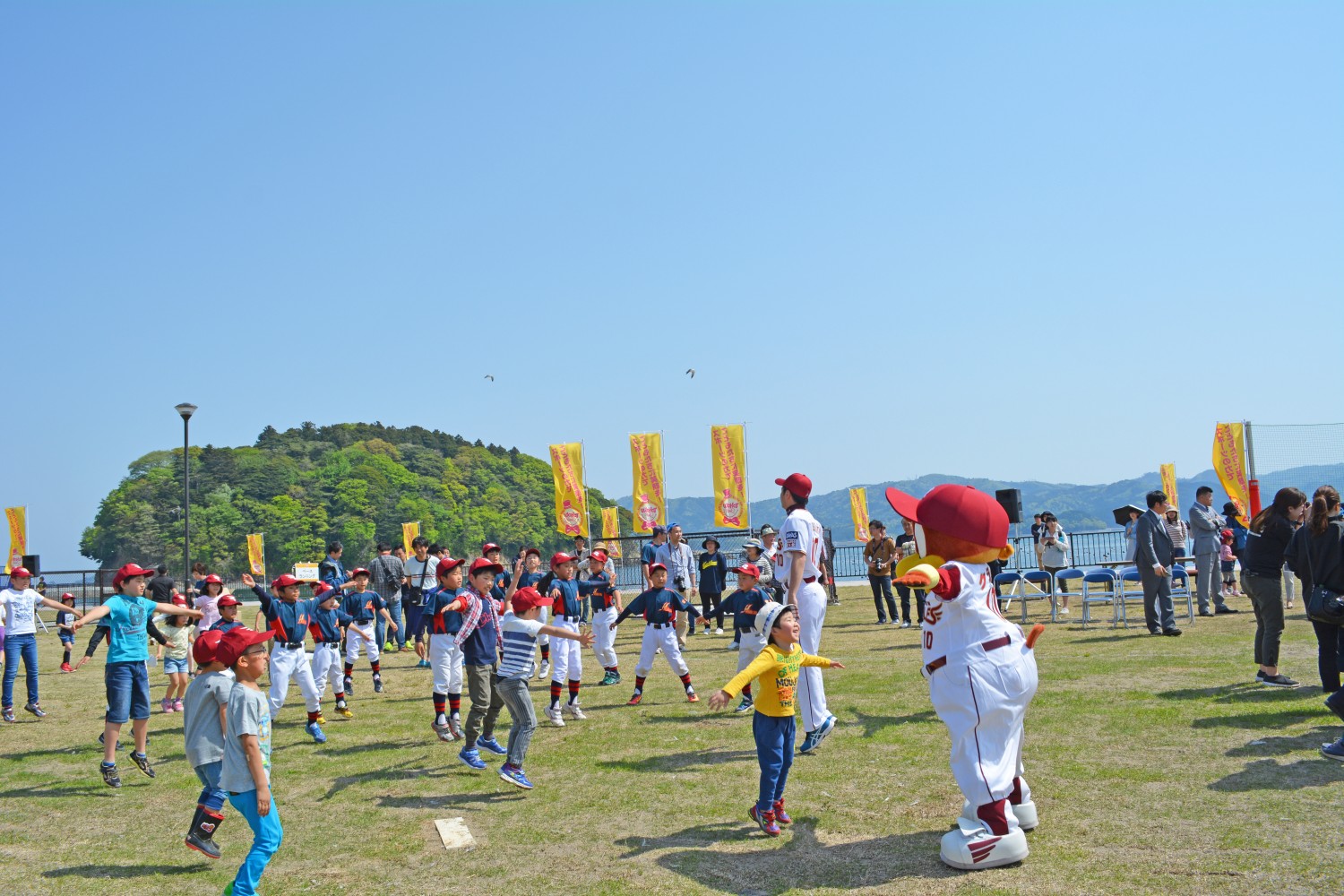 屋外の芝生広場で、野球帽をかぶった子どもたちがプロ野球選手やマスコットキャラクターと一緒に両腕を外に広げて元気よく飛び跳ねている写真