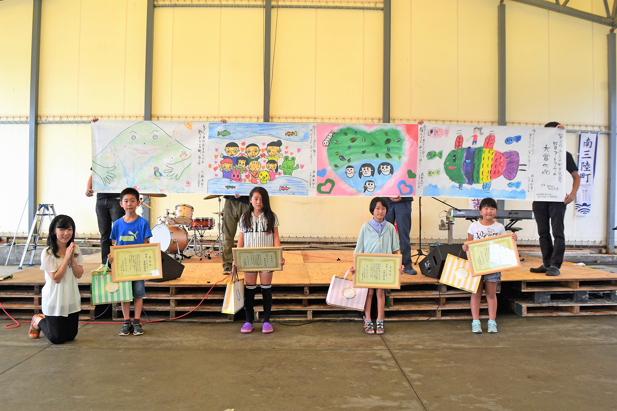 4人の子供達が賞状とプレゼント袋を持ってステージの前に並び、後ろに色鮮やかな受賞作品が掲げられている写真