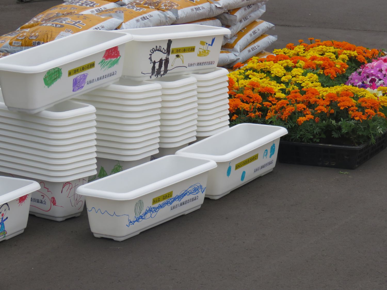 土の袋とオレンジ・黄色・ピンクの花の苗が並べられ、手前に子どもたちの絵や文字が描かれた大量の白いプランターが積み重ねられている写真