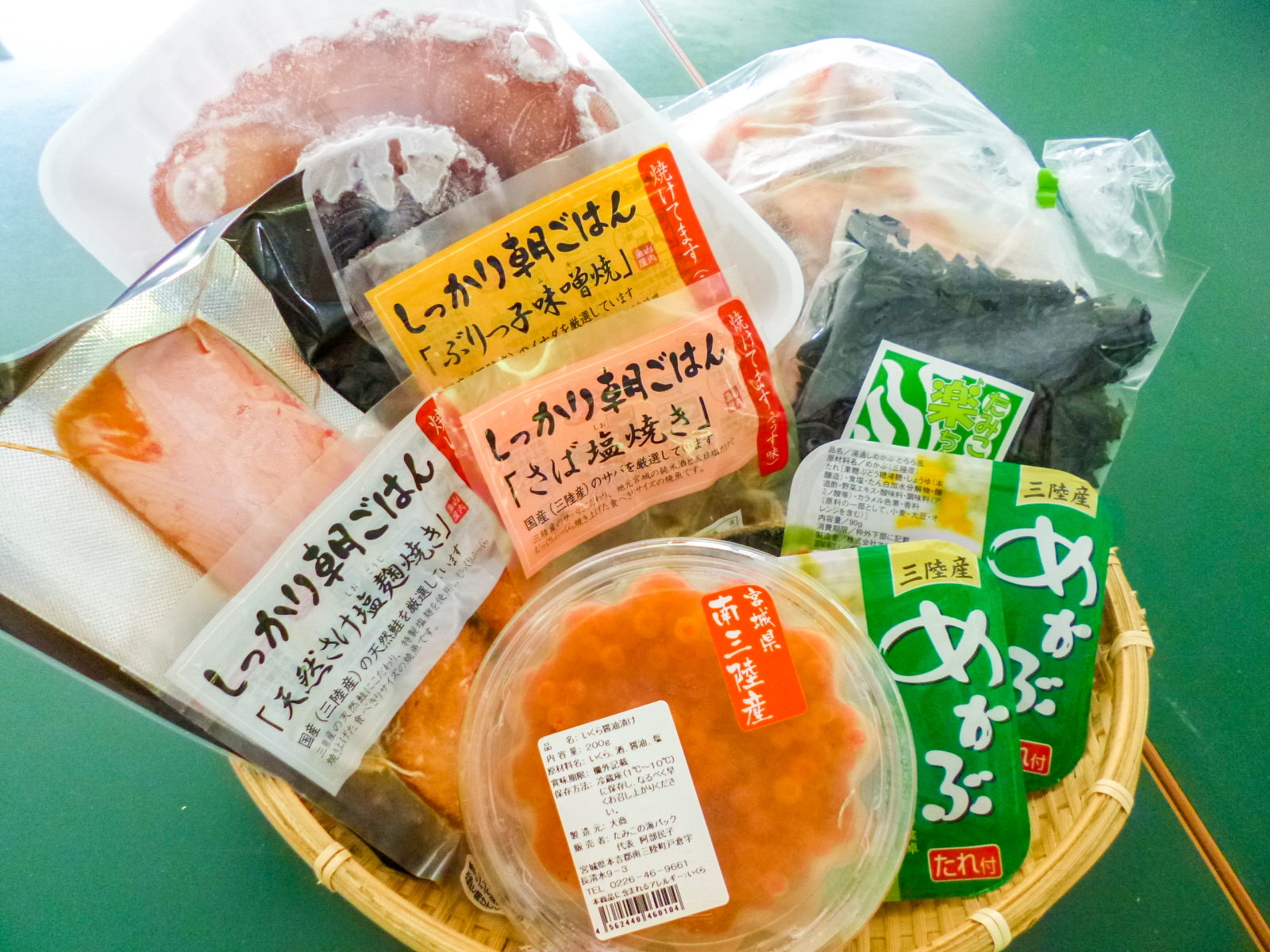 南三陸町産のめかぶやいくら、真空パックに入った焼き魚、乾燥わかめなどの海産物加工品がかごの中に詰め合わせられている写真