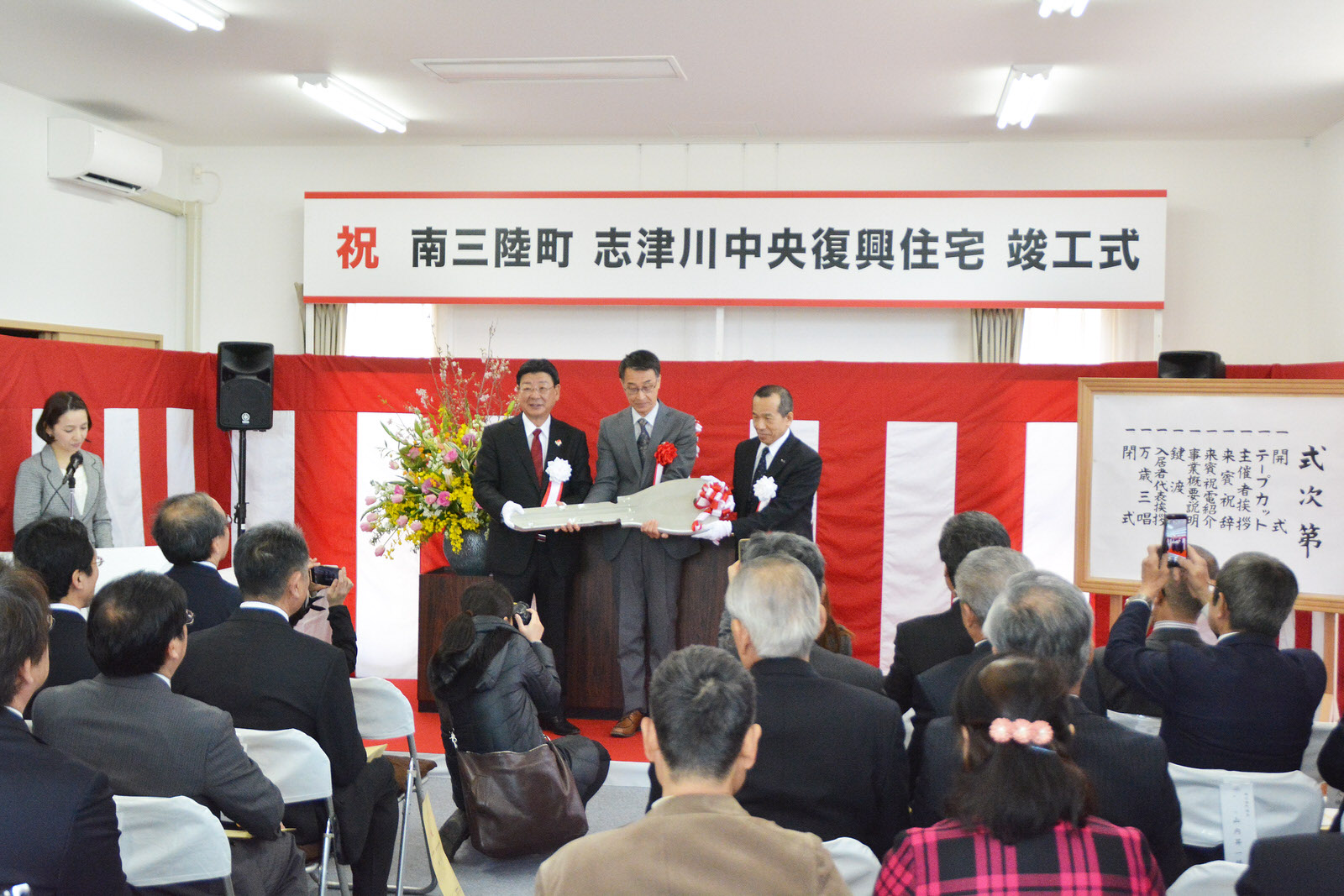 「祝 南三陸町志津川中央復興住宅の竣工式」と書かれた横断幕が掲げられ、紅白幕が張られた室内で壇上に立った男性3人が大きなカギのレプリカを持って立っている写真