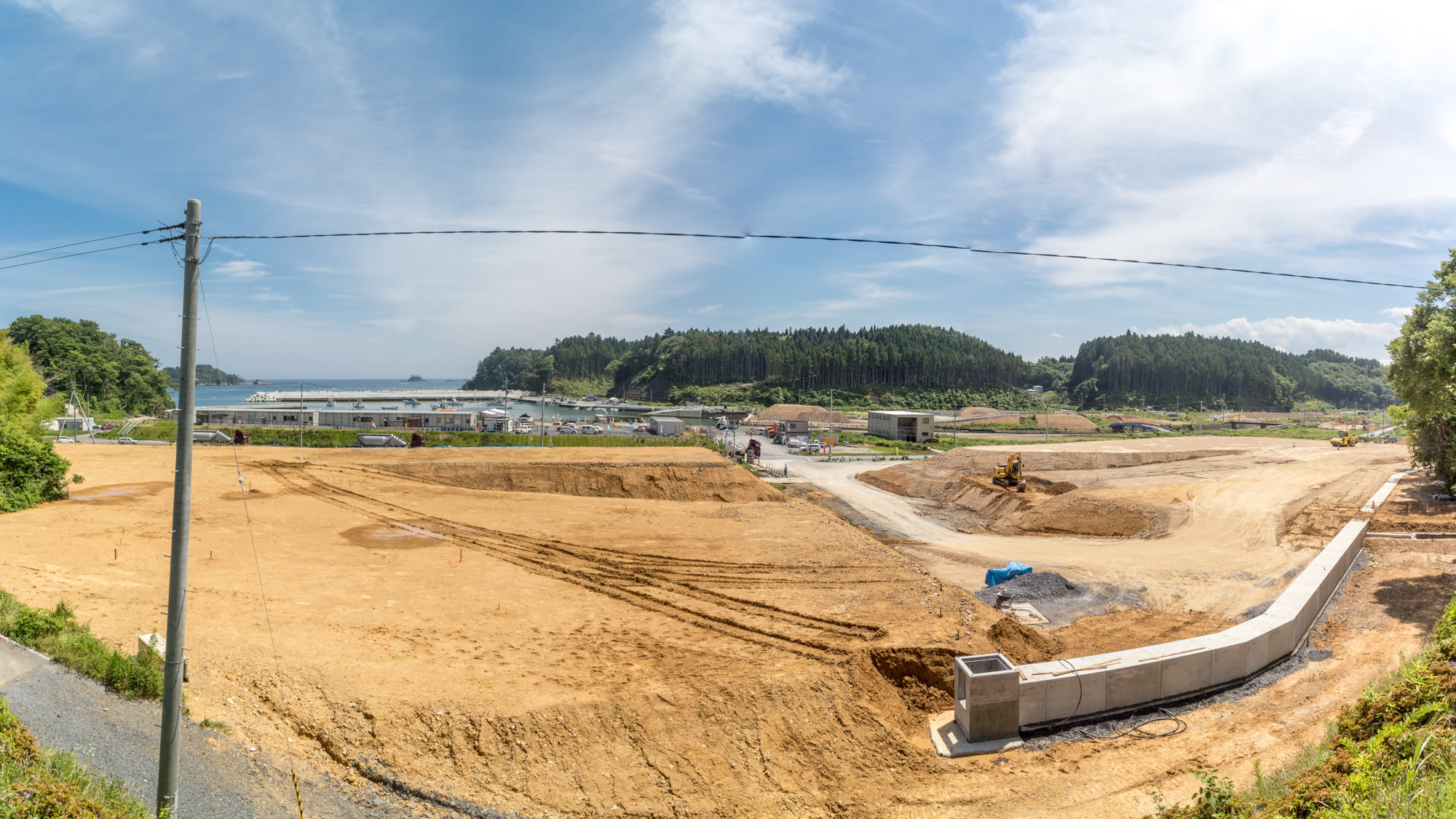 海に面した造成中の土地の定点カメラによる風景で広大な更地が広がっており、黄土色の土がむき出しのまま整地され、地面には重機のタイヤ跡が残り、建設用資材が置かれた状態の 基礎工事の跡を写した写真