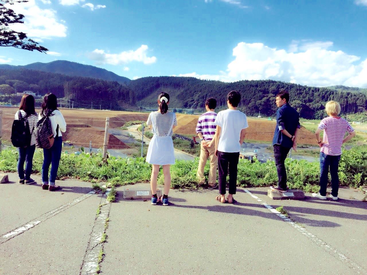 男女の若者たちが高台の駐車場に立ち、山の前に広がる造成地を見つめている後ろ姿の写真