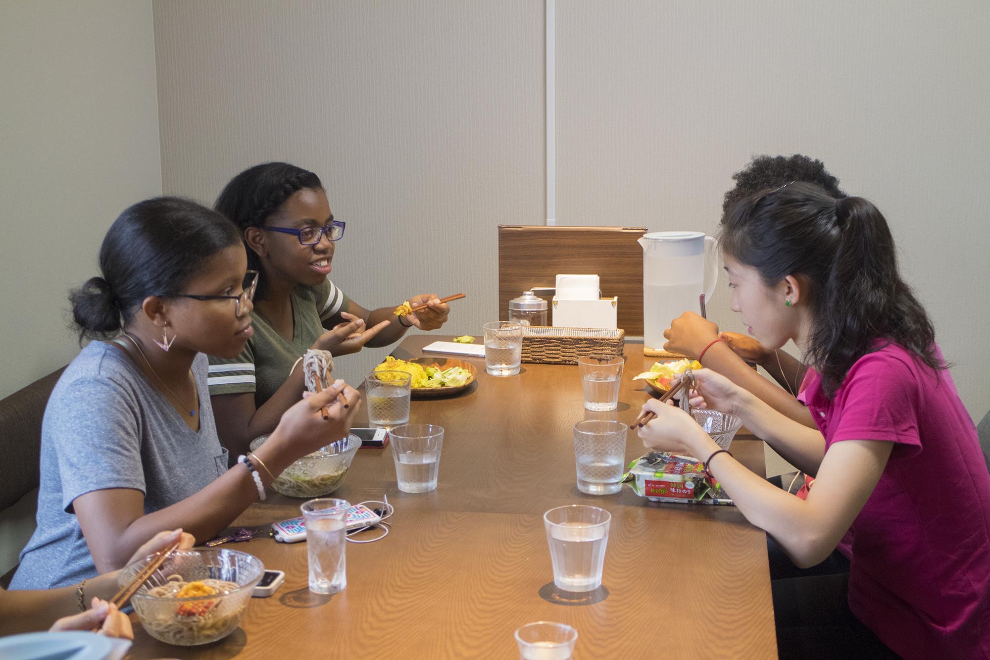 4人のアメリカ人の女子学生たちが、テーブルを囲んで料理を箸で食べながら楽しそうに会話をしている写真