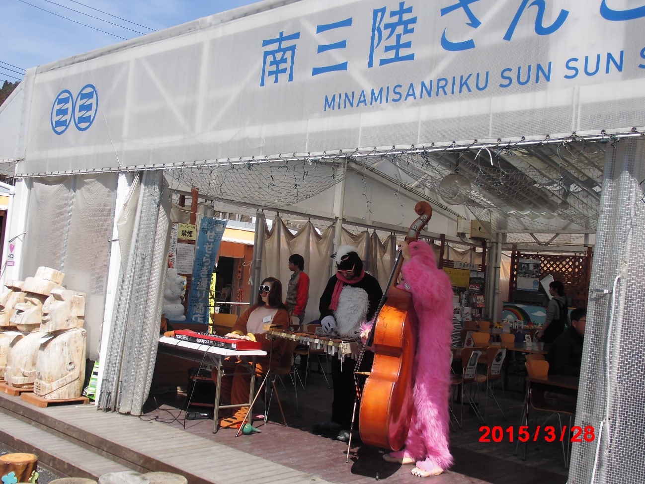 南三陸さんさん商店街の前で、動物の着ぐるみを着た数人の人物が並んで楽器を演奏している様子が写っている写真
