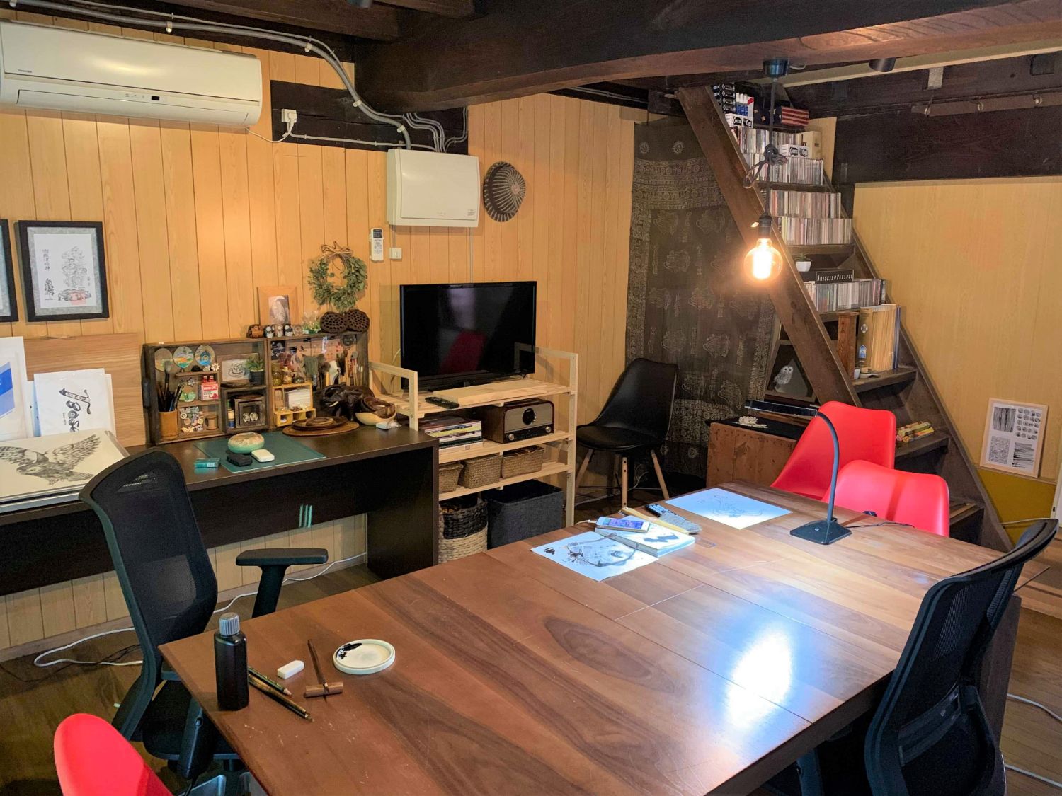 木の大きなテーブルが置かれ、赤と黒の椅子やテレビ、壁際の棚に画材や飾りが整然と並び、天井の梁が見えている木目の壁に囲まれた作業部屋の写真